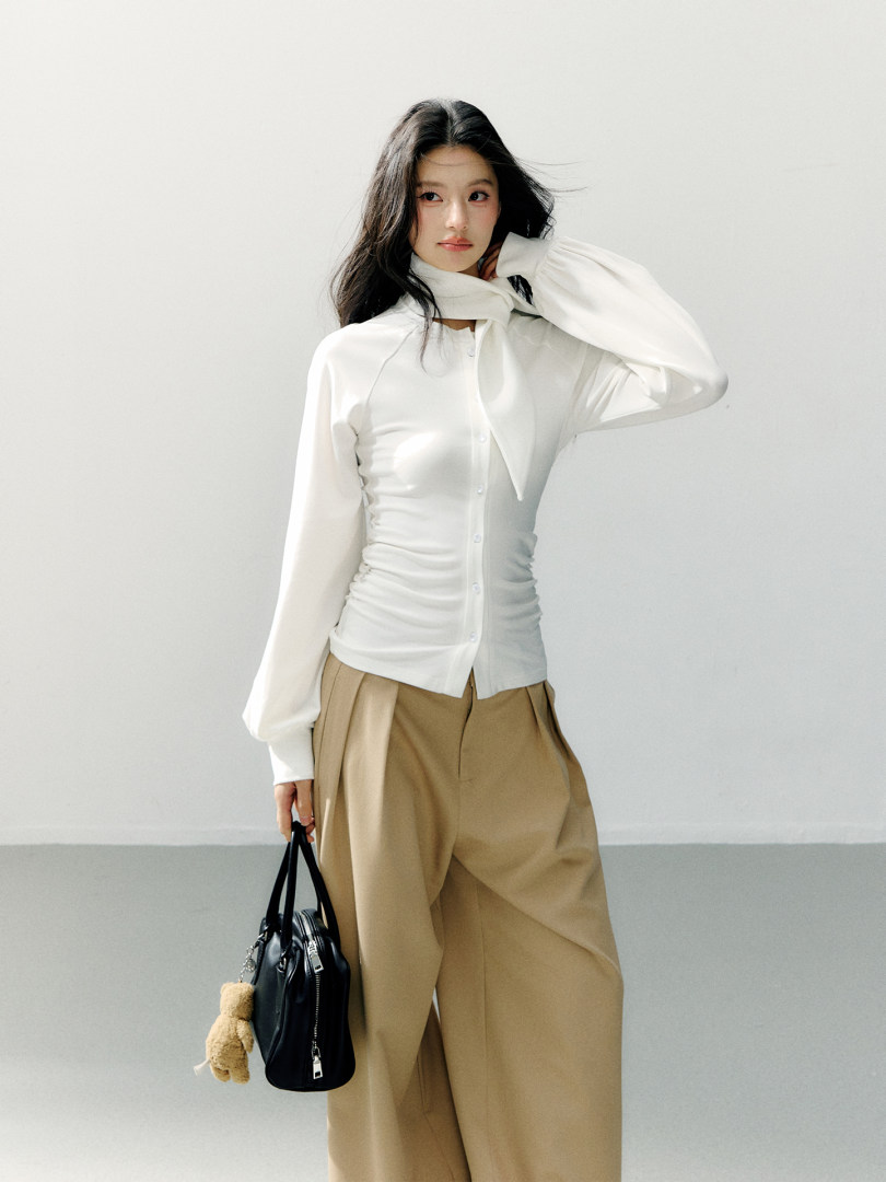 Model wearing a white fitted blouse with a scarf detail and beige wide-leg trousers, holding a black handbag