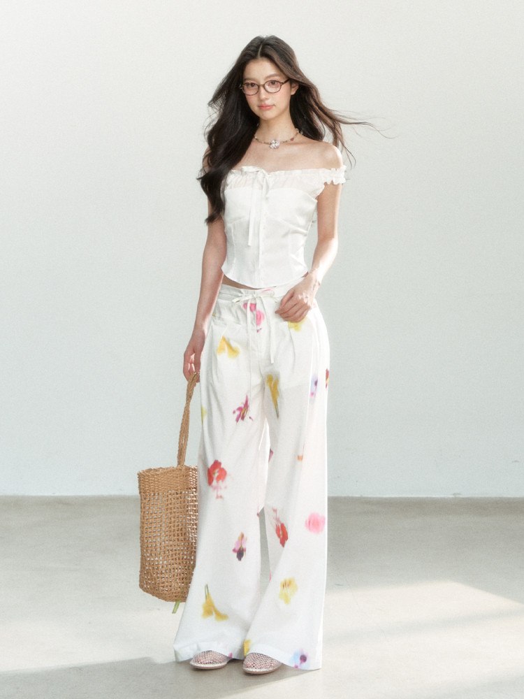 Model wearing a white off-shoulder top with floral wide-leg pants and a woven tote bag