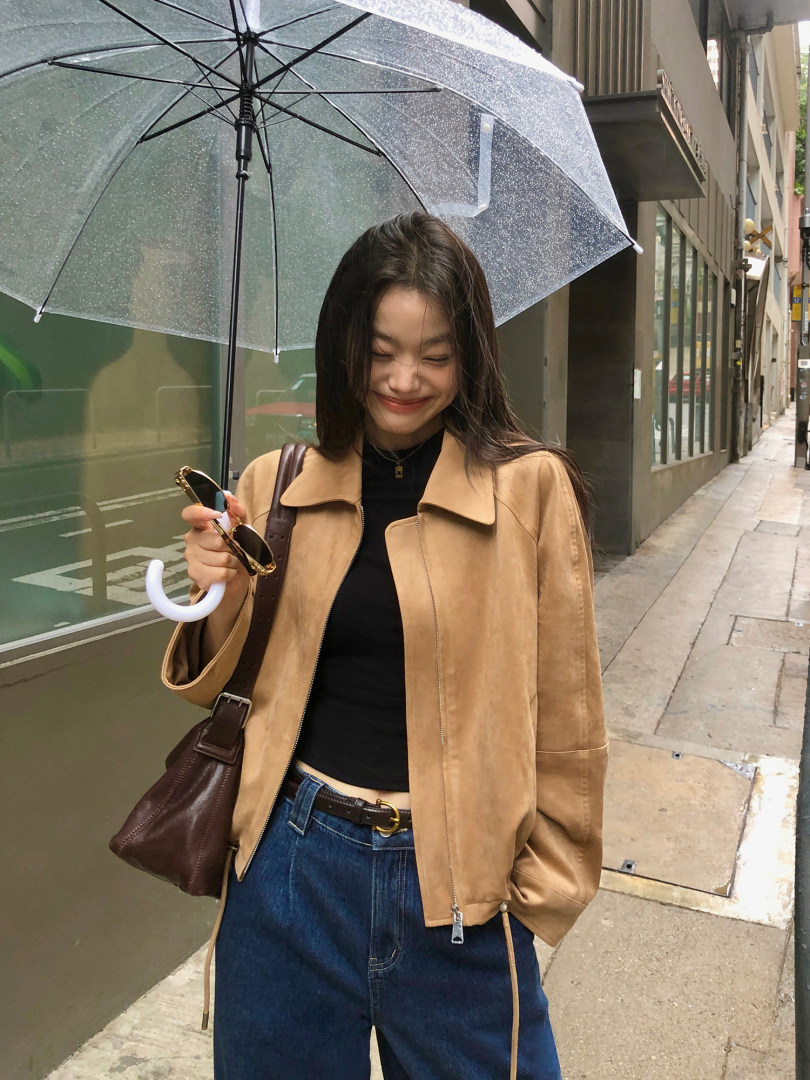 Woman wearing a tan jacket with jeans and a brown shoulder bag under a clear umbrella
