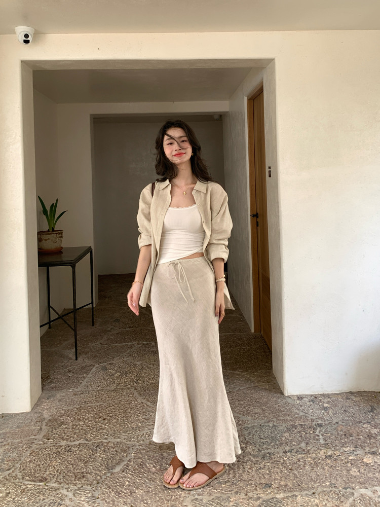 Woman wearing a beige button-up shirt and matching long skirt with a white tank top and sandals