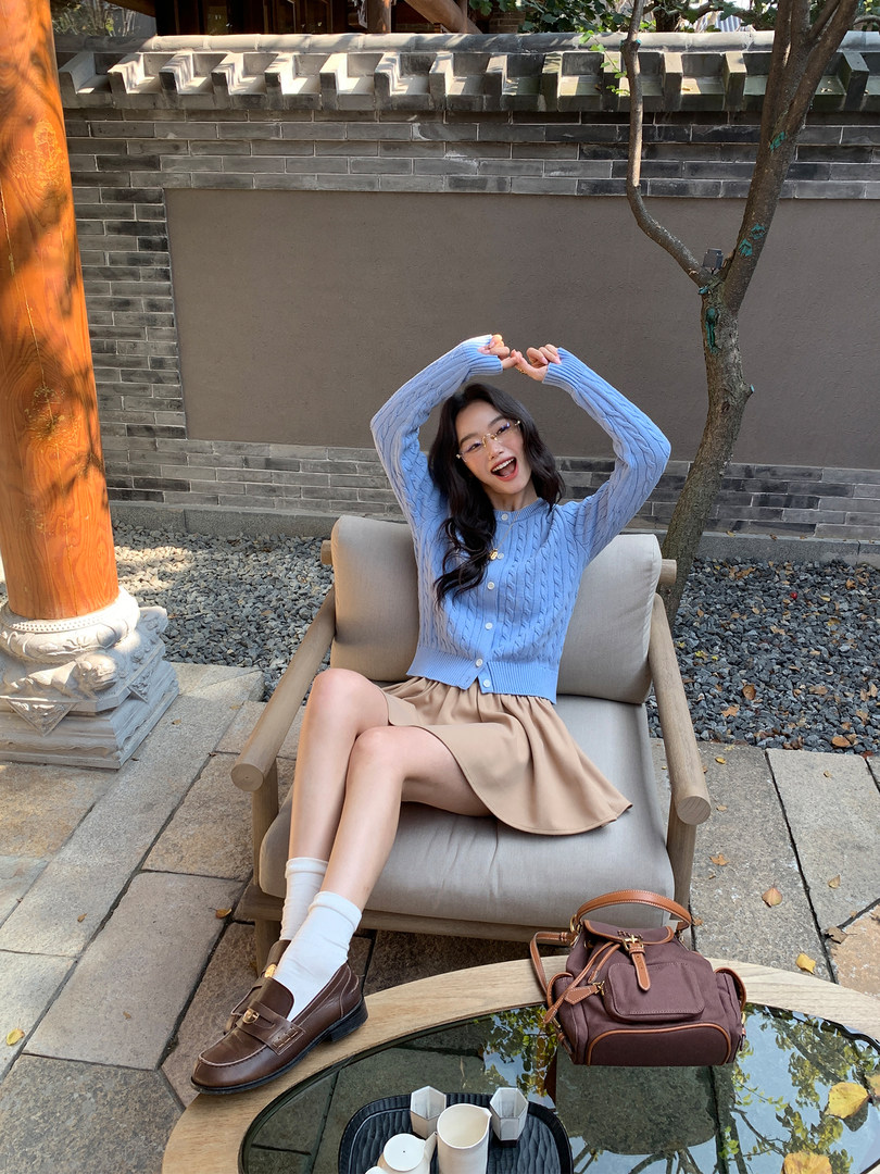 Model wearing a light blue cable-knit cardigan with a beige skirt and brown loafers outdoors