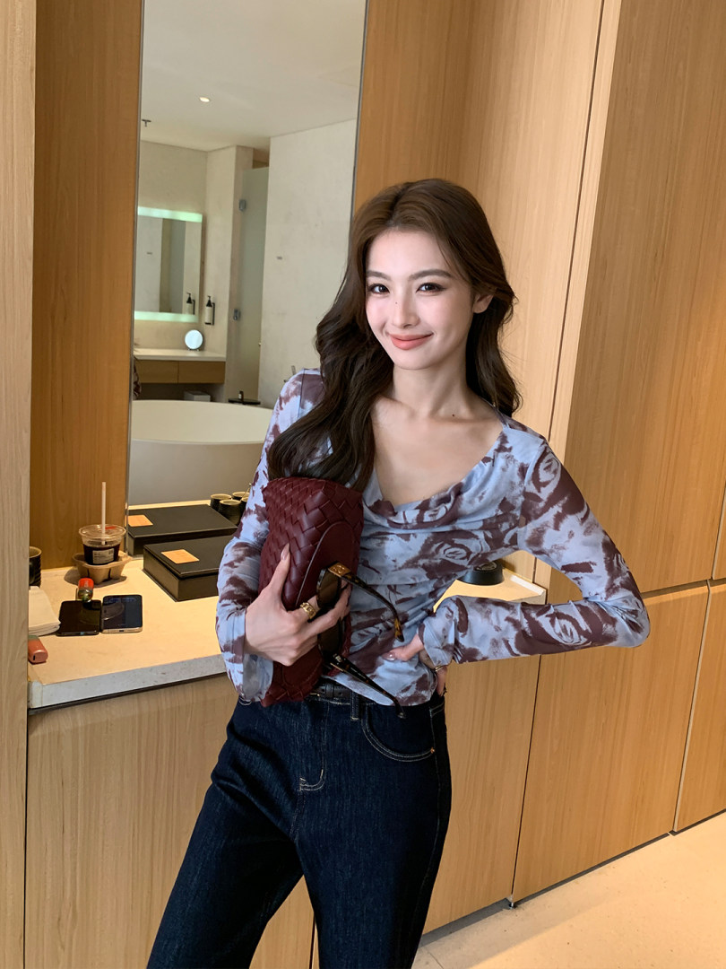 Woman wearing a patterned long-sleeve top and dark jeans while holding a woven clutch