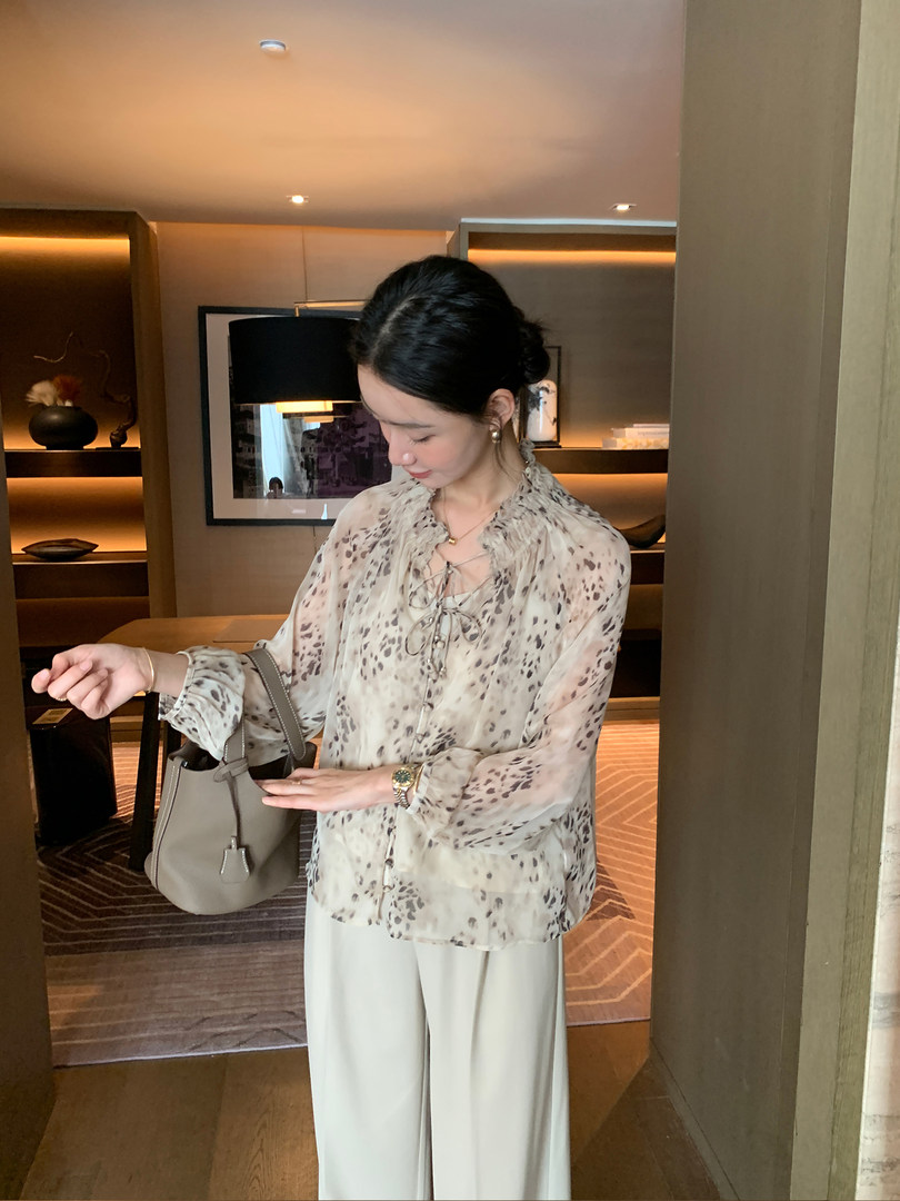 Woman wearing a sheer patterned blouse and wide-leg trousers with a taupe handbag