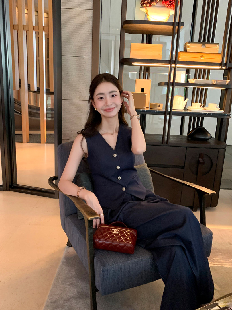 Woman wearing a navy sleeveless buttoned outfit while seated indoors with a small red quilted handbag