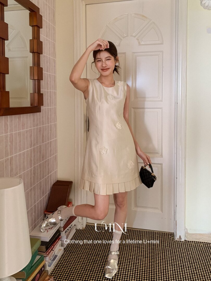 Model wearing a cream sleeveless mini dress with floral embellishments and a pleated hem