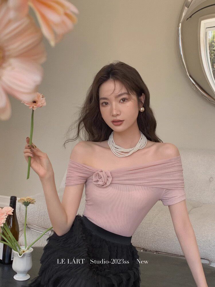 Model wearing a pink off-shoulder top with floral detail and pearl necklace