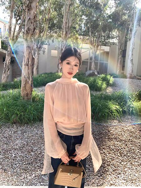 Woman wearing a sheer blush long-sleeve blouse and holding a beige handbag outdoors