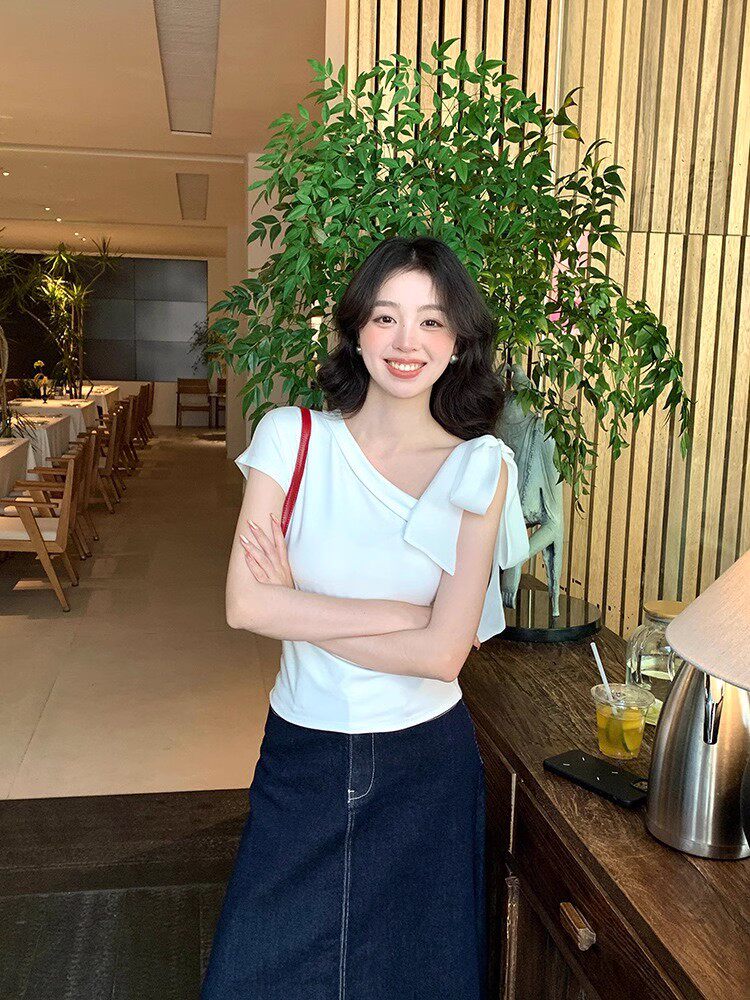 Woman wearing a white one-shoulder top with a bow and a dark denim skirt in a restaurant