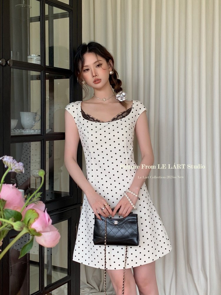 Model wearing a white polka dot sleeveless dress and holding a black quilted handbag