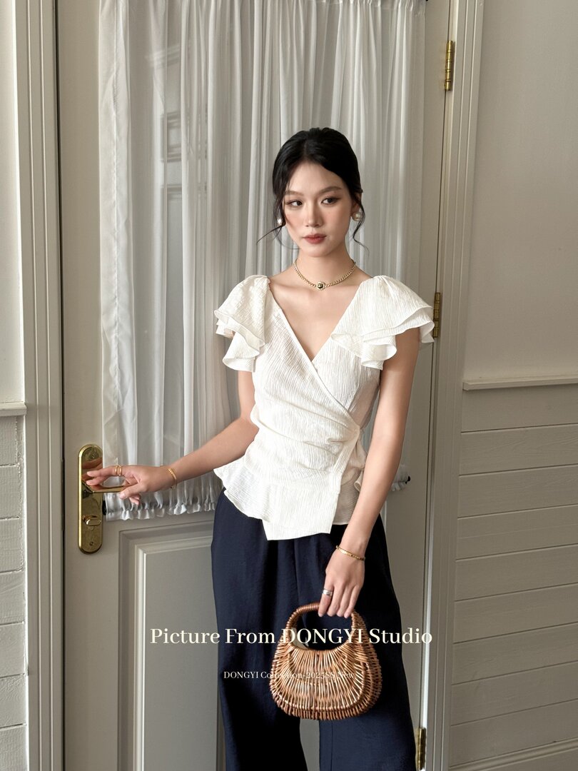 Model wearing a white ruffled wrap blouse with navy pants and a wicker handbag