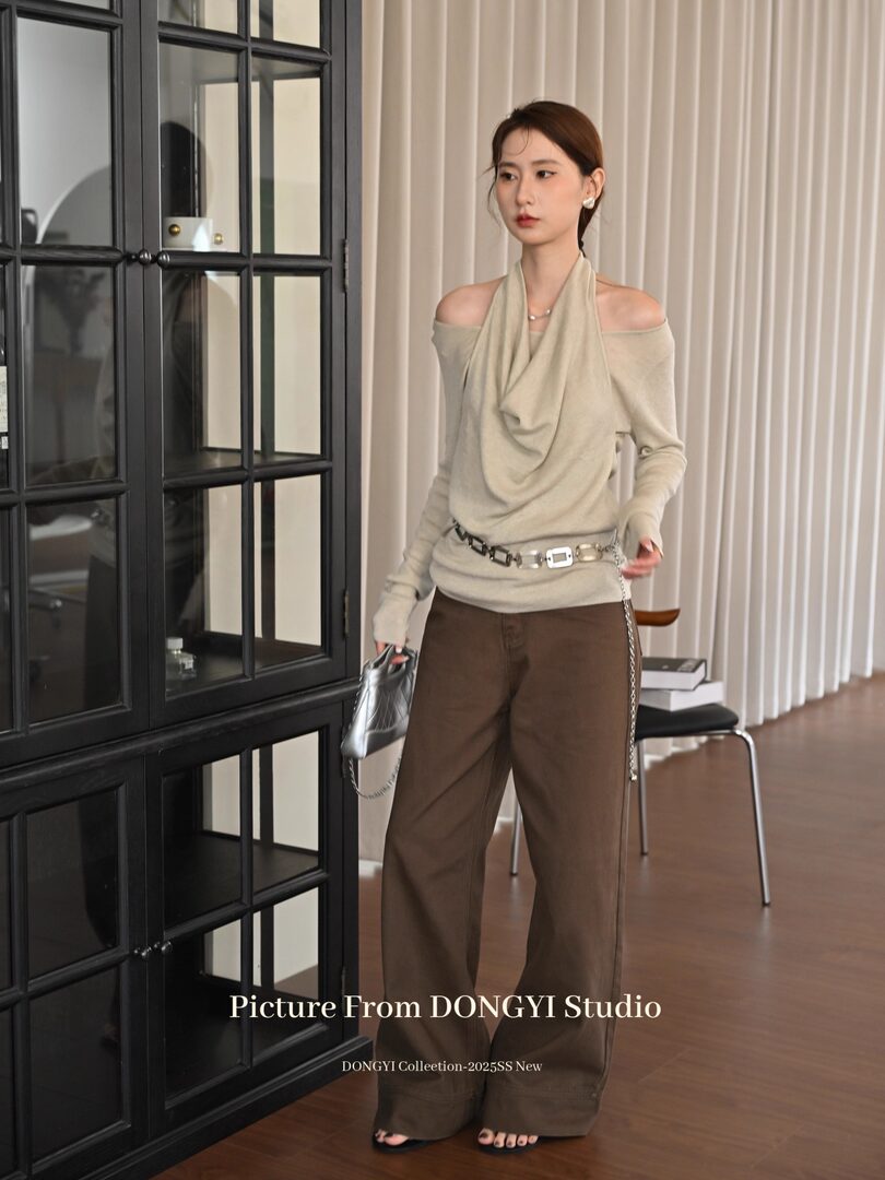 Model wearing a beige off-shoulder draped top with brown wide-leg pants and a chain belt