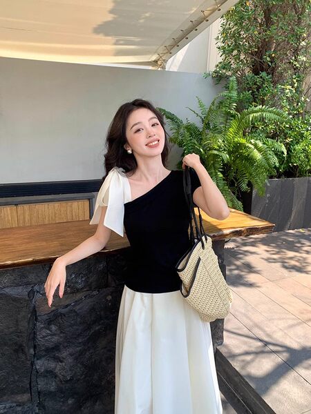 Woman wearing a black one-shoulder top with a white skirt and carrying a woven bag outdoors