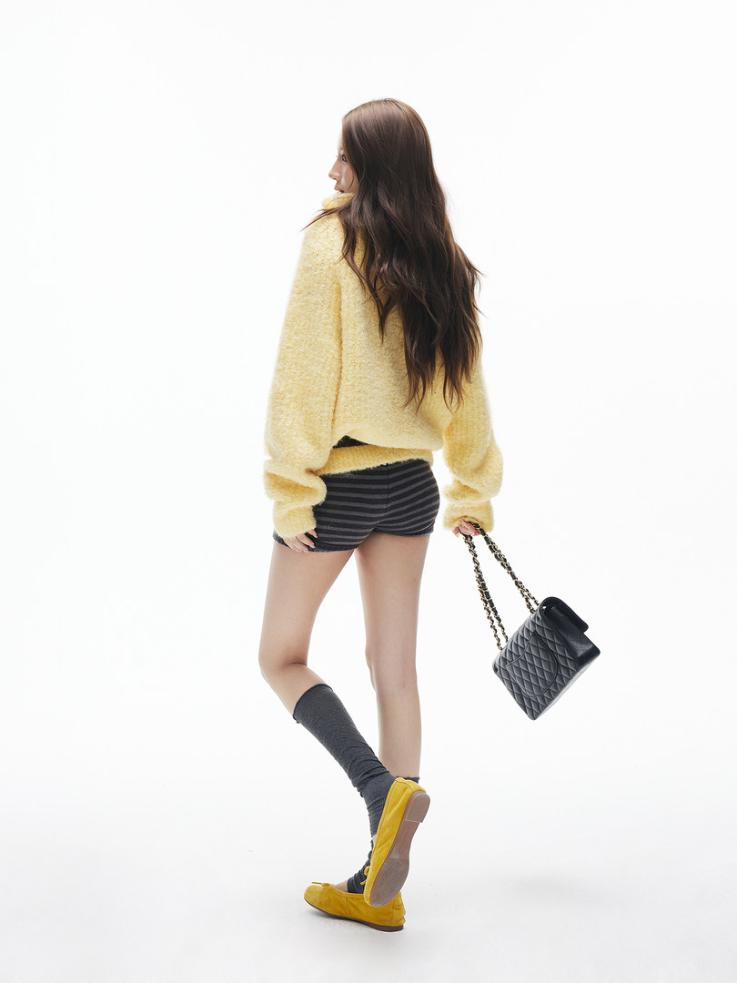 Model wearing a pale yellow fuzzy sweater with striped shorts, socks, yellow flats, and a black quilted handbag