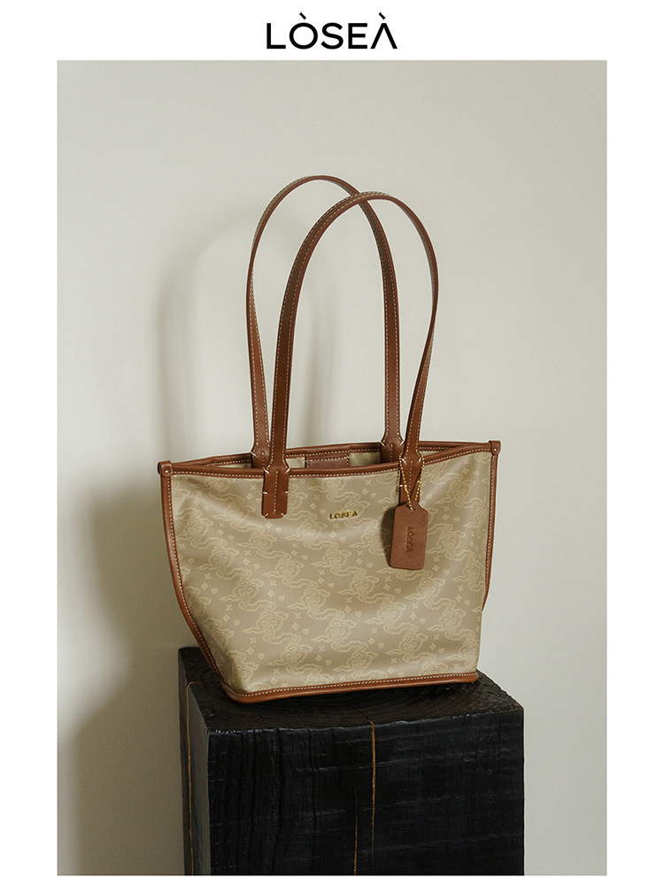 Beige monogram tote bag with brown leather handles on a pedestal