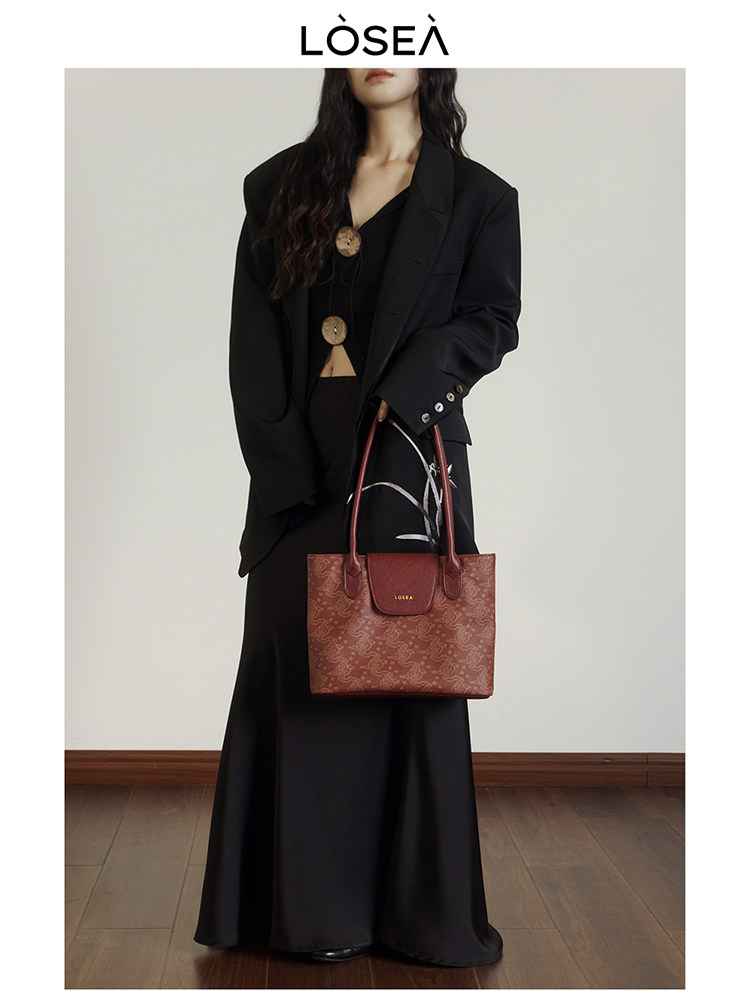 Model wearing a black blazer and long skirt while holding a patterned brown tote bag