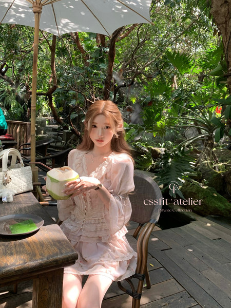 A model wearing a light pink lace dress seated outdoors with a coconut drink