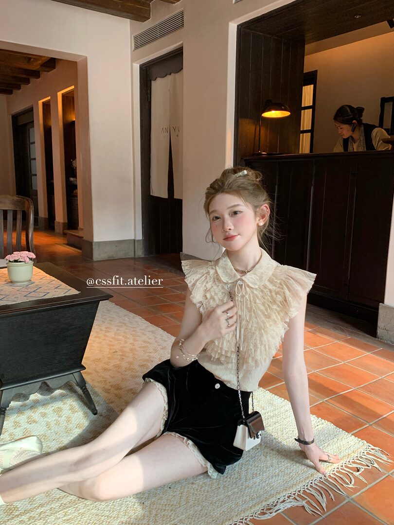Model wearing a cream ruffled sleeveless blouse with black shorts in a warm indoor setting