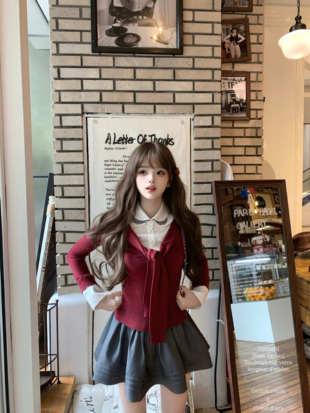 Model wearing a red knit cardigan over a white collared shirt with a gray skirt in a cozy indoor setting