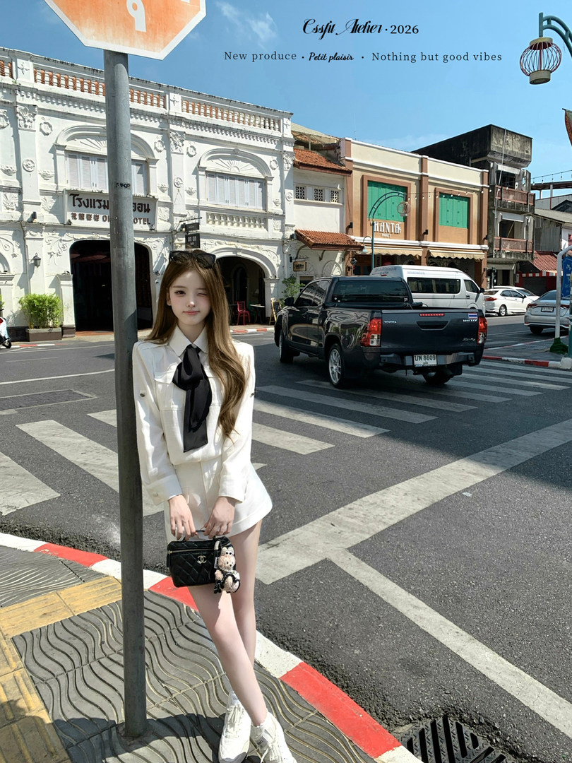 Woman modeling a white preppy outfit with black bow tie and small black handbag on a city street