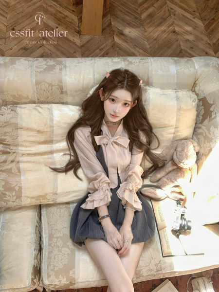 Model wearing a light pink bow blouse with a pleated dark skirt while seated on a sofa