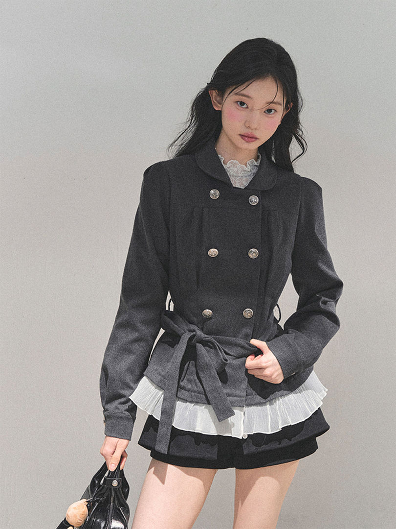 Model wearing a dark gray double-breasted belted jacket with a layered white ruffle hem skirt