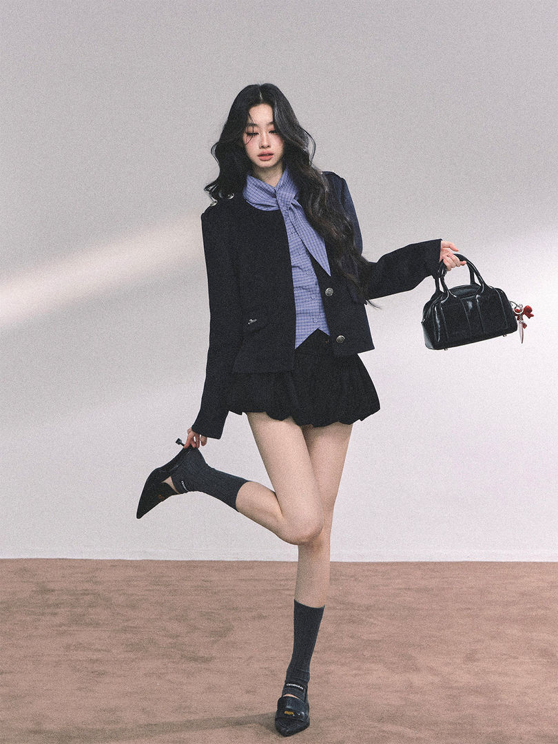 Model wearing a black blazer outfit with a pleated skirt and holding a black handbag