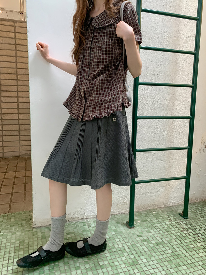 Model wearing a brown plaid short-sleeve top, gray pleated skirt, and black Mary Jane shoes
