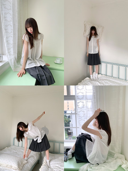 Model wearing a white polka dot blouse with a black pleated skirt in a bedroom setting