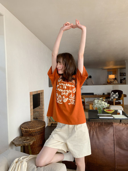 Woman wearing an orange graphic oversized T-shirt with cream shorts in a casual indoor setting