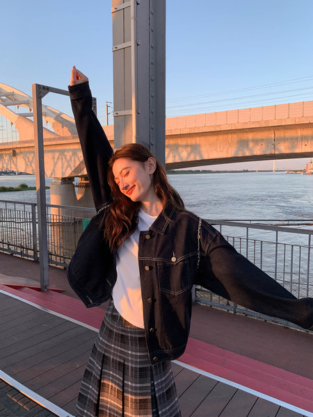 Woman wearing a black denim jacket over a white top with a plaid skirt outdoors by the water
