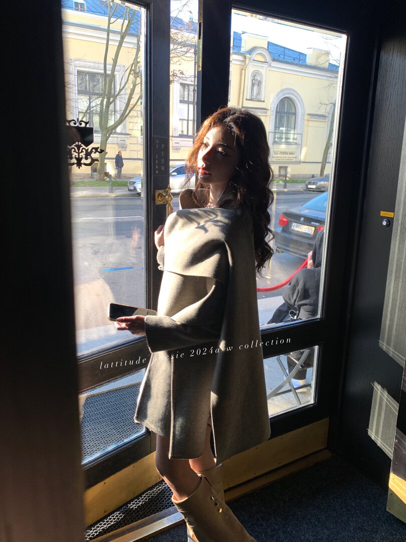 Model wearing a taupe oversized cape coat indoors by a window