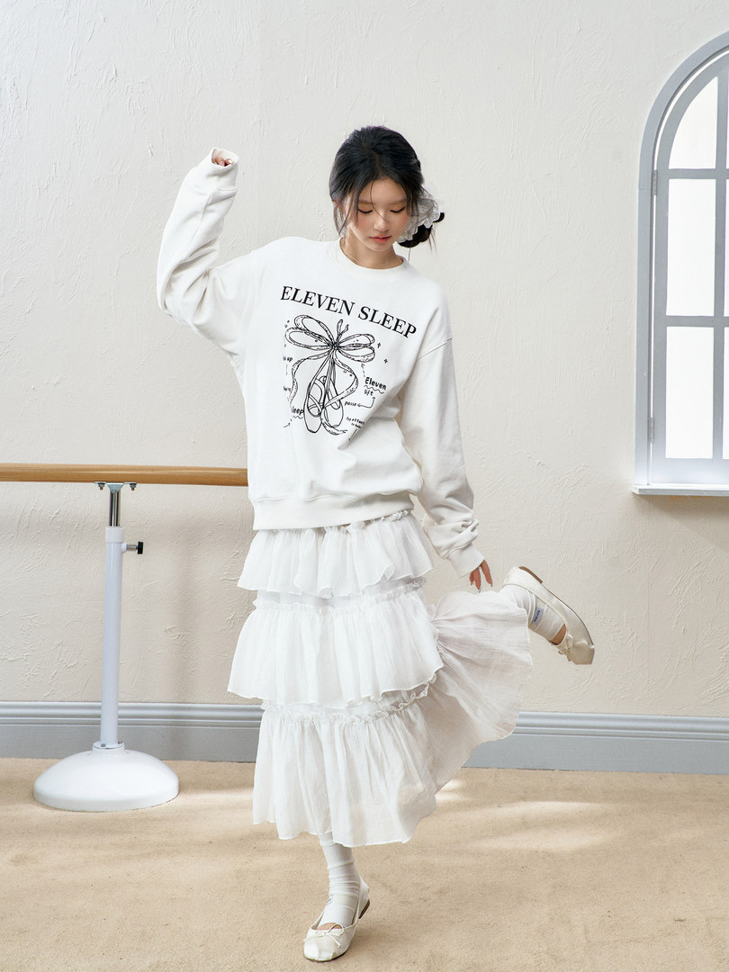 Model wearing a white graphic sweatshirt with a tiered white skirt and white flats