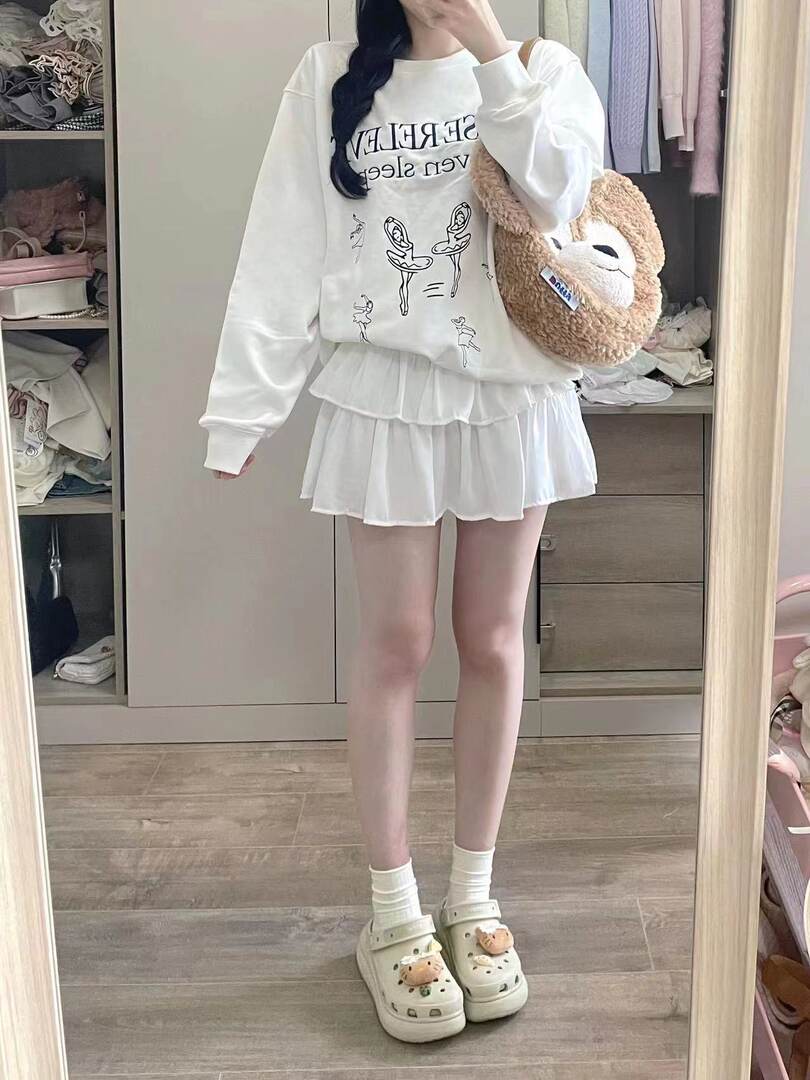 Woman wearing a white graphic sweatshirt, pleated mini skirt, and platform clogs in a mirror selfie
