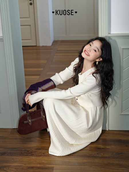 Model wearing a white long-sleeve dress while sitting with a handbag