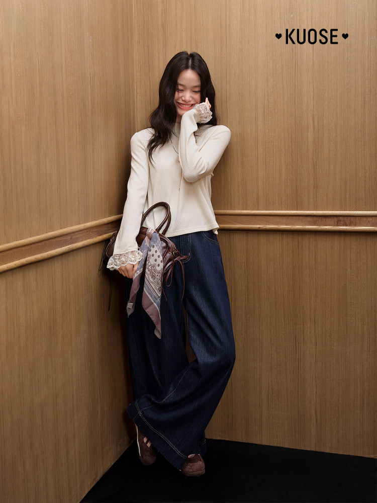Woman wearing a cream long-sleeve top with lace cuffs and wide-leg jeans, holding a handbag