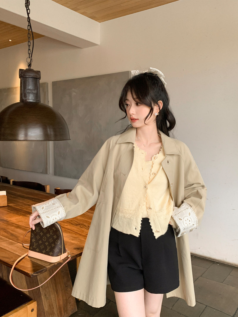Woman wearing a beige trench coat over a cream cardigan and black shorts, holding a monogram handbag