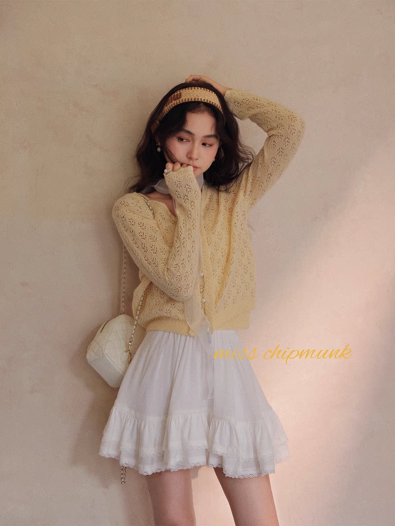 Model wearing a light yellow knit cardigan with a white ruffled skirt and quilted shoulder bag