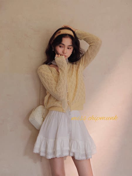 Model wearing a light yellow knit cardigan with a white ruffled skirt and quilted shoulder bag
