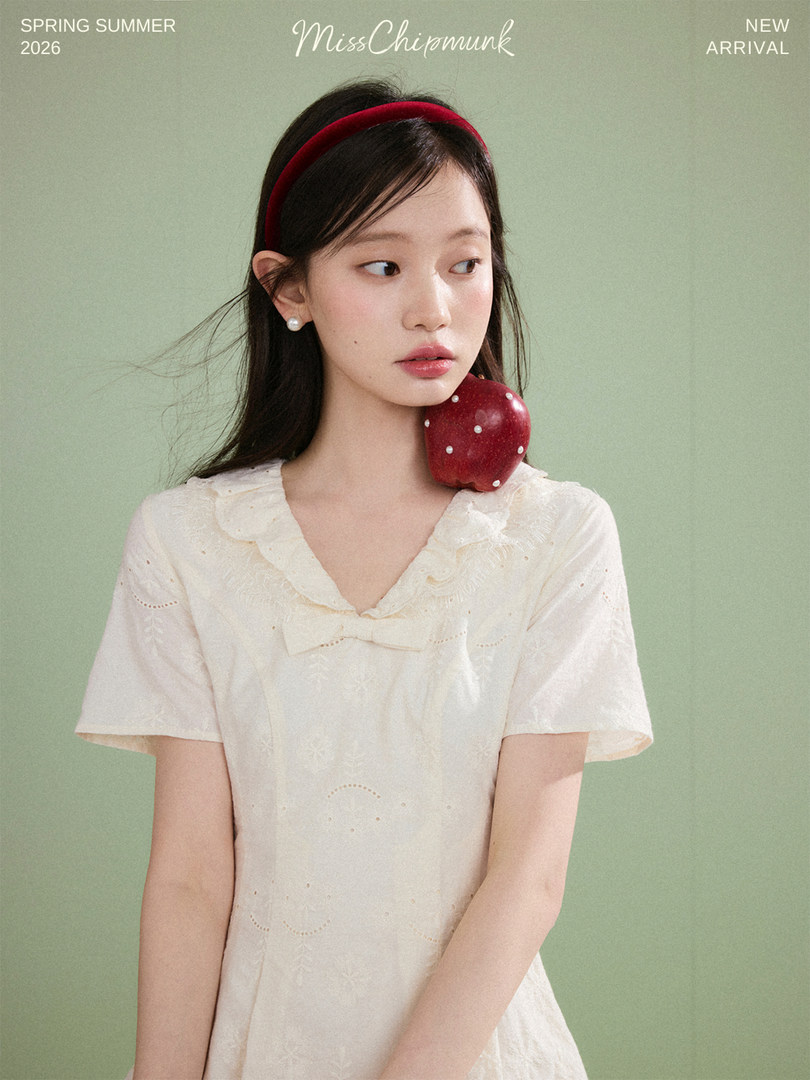 Model wearing a cream short-sleeve dress with a red headband and apple accessory against a green background
