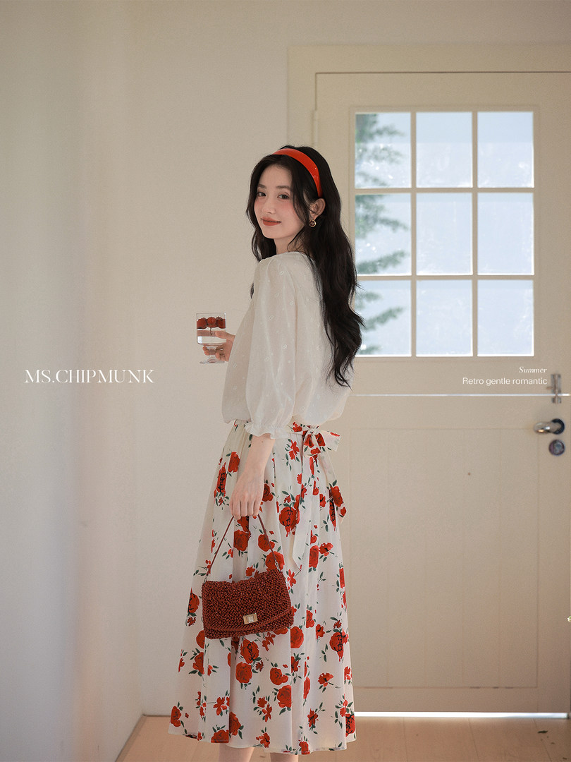 Woman modeling a white blouse and red floral skirt with a small woven handbag indoors