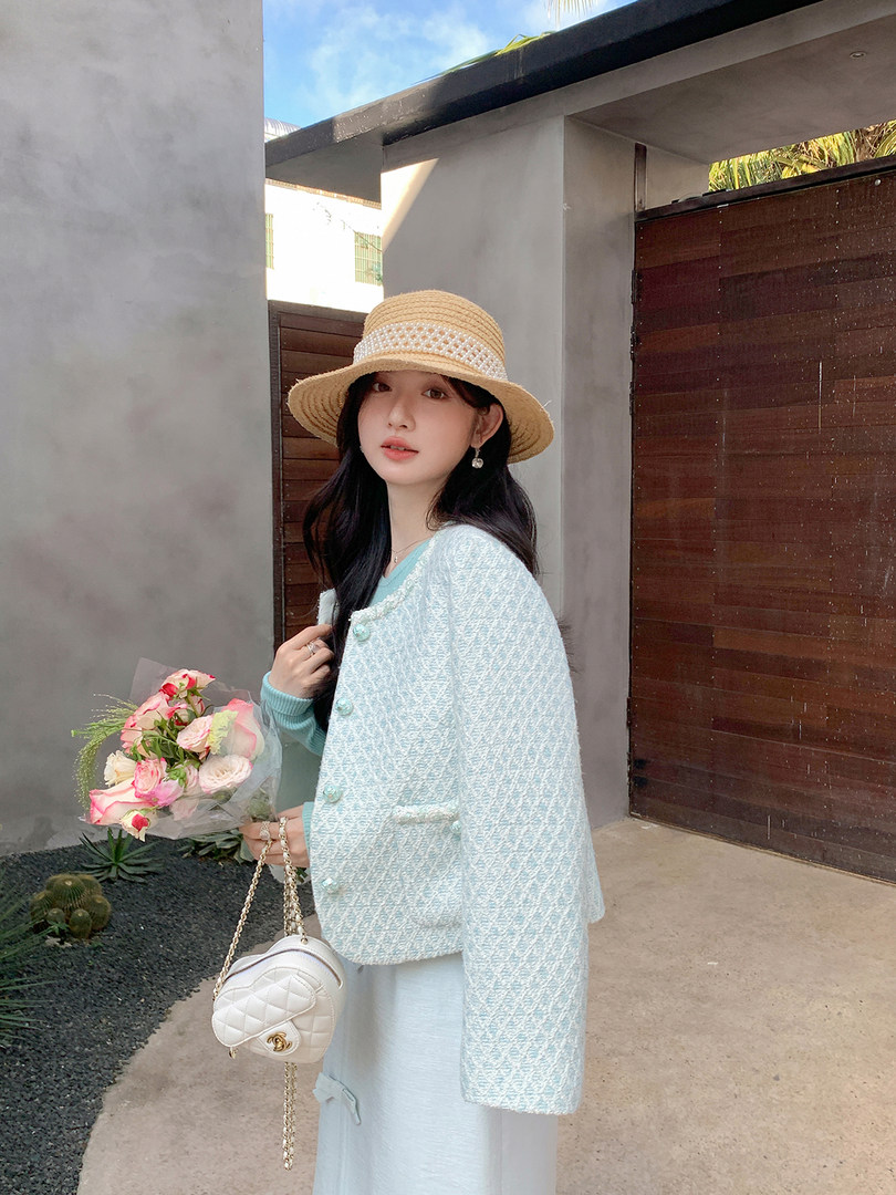 Woman modeling a light blue textured jacket and straw hat while holding a quilted white shoulder bag