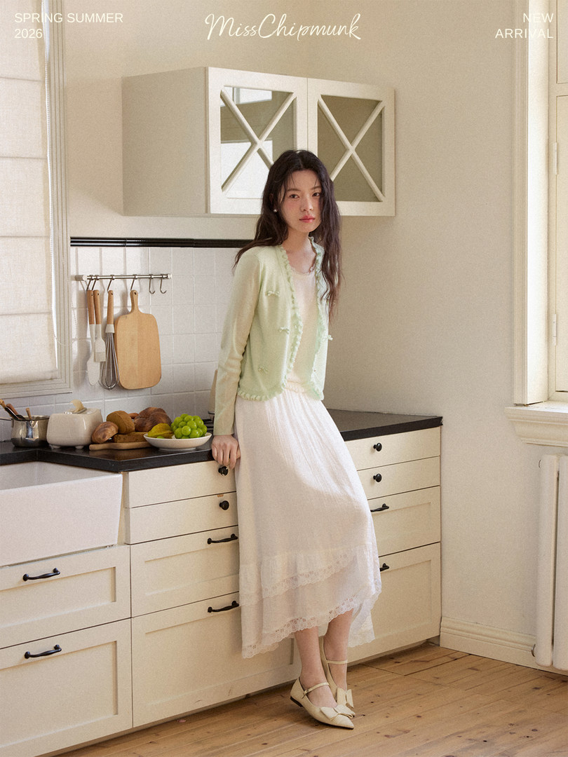 Woman modeling a light green cardigan and white skirt in a kitchen setting with promotional text overlay