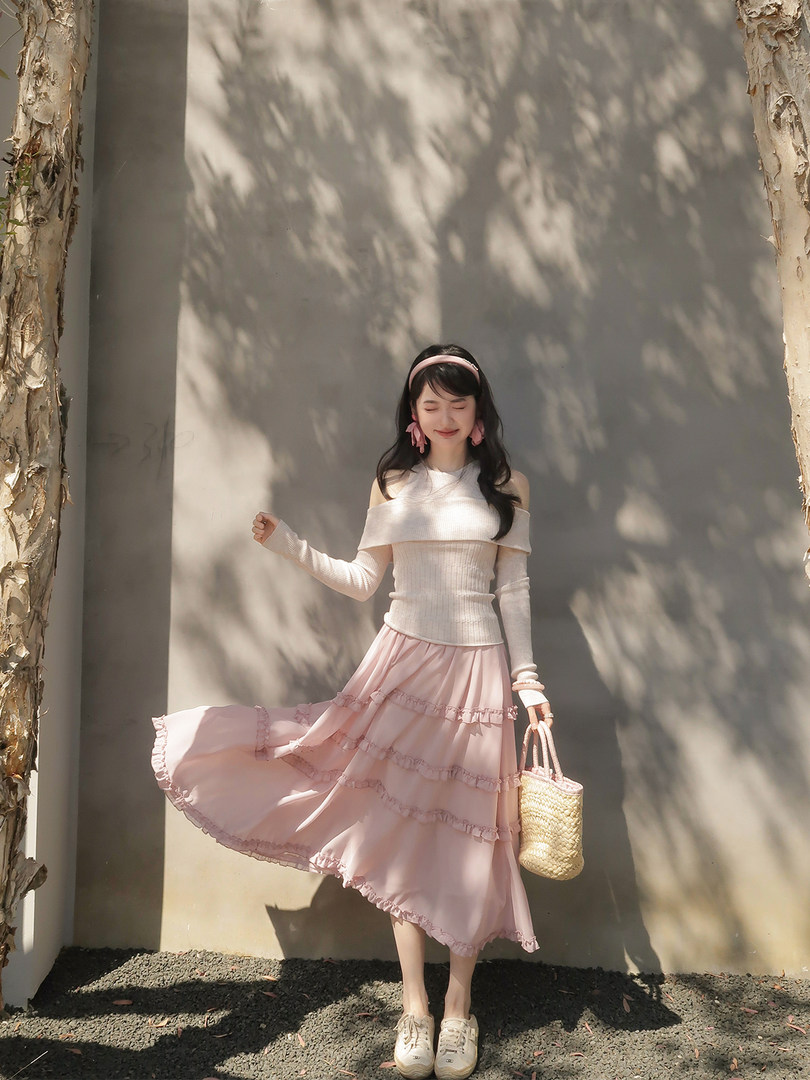 Woman modeling a white off-shoulder top with a pink ruffled skirt and straw handbag