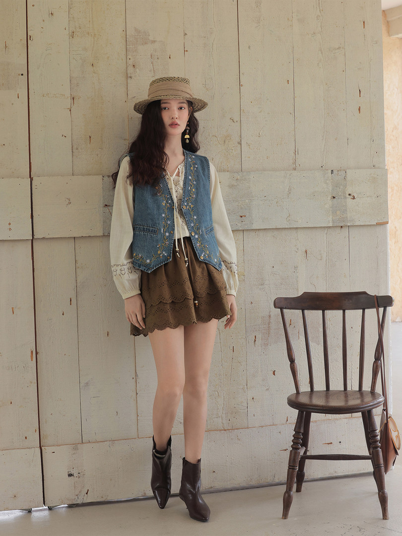 Fashion model wearing a boho-inspired outfit with denim vest, blouse, skirt, hat, and boots