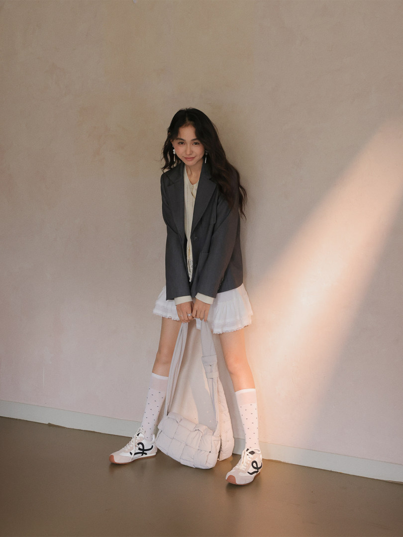 Woman modeling a casual fashion outfit with blazer, white skirt, sneakers, and shoulder bag