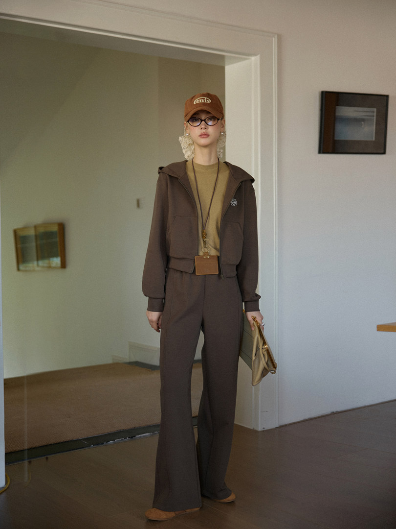 Woman wearing a brown casual hoodie and matching pants with a cap and handbag indoors