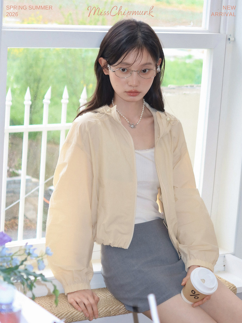 Model wearing a beige lightweight jacket over a white top and gray skirt