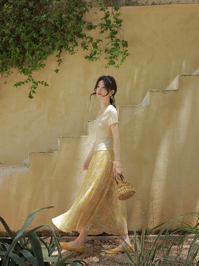Woman wearing a light cream top and yellow floral pleated skirt outdoors with a small woven handbag