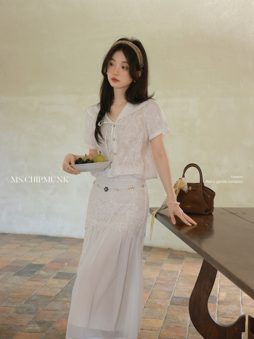 Model wearing a white lace two-piece outfit with a small brown handbag in a styled studio setting