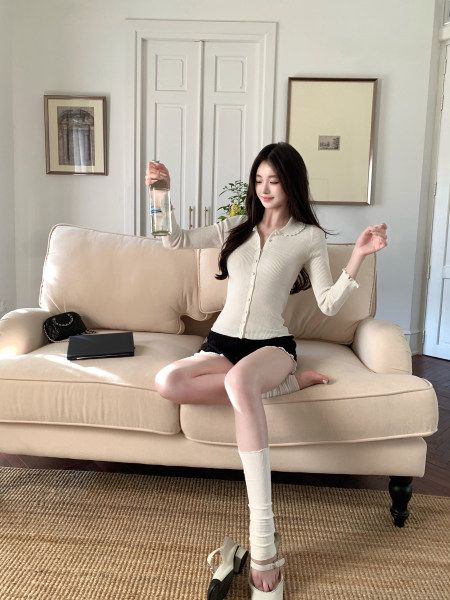 Woman sitting on a beige couch wearing a cream ribbed cardigan, black shorts, and white knee-high socks with white sandals.